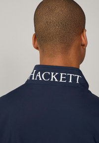 Marineblå jakke med høje krave, glat tekstur og hvid "HACKETT" branding på ryggen. Enkel og stilren design.