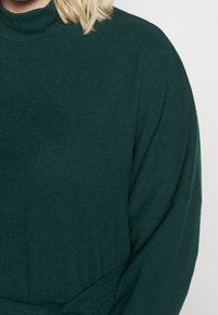 Maglione a maniche lunghe e colletto alto di colore verde scuro, realizzato in tessuto morbido con una vestibilità rilassata. Caratterizzato dalla sua texture liscia e dal design minimale.
