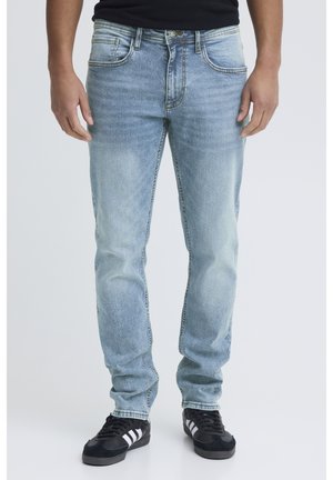 Person trägt hellblaue ausgewaschene Jeans und schwarze Turnschuhe mit weißen Streifen, steht vor einem schlichten hellen Hintergrund.