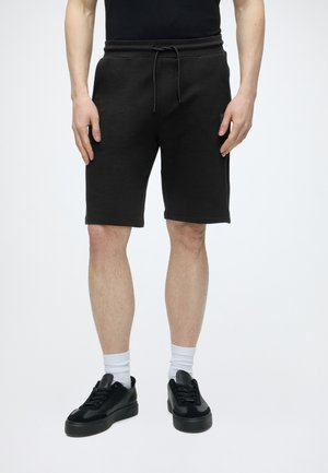 Uomo che indossa pantaloncini neri con coulisse, scarpe da ginnastica nere, calzini bianchi e una maglietta nera, in piedi contro uno sfondo bianco uniforme.