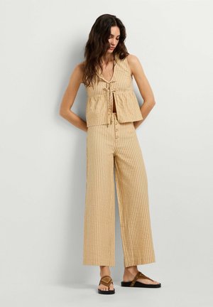 Mujer vistiendo un top sin mangas beige a rayas con lazos delanteros y pantalones de pierna ancha a juego con detalles de botones, de pie frente a un fondo liso.