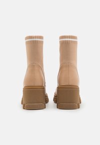 Botas estilo sneaker en color marrón claro con un upper de punto elástico, suela de goma gruesa y tacón texturizado. Presentan detalles en blanco a rayas en la parte superior.