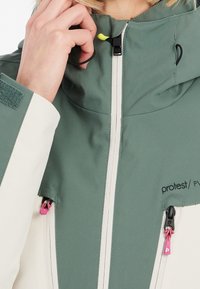 Veste verte et crème avec un col montant. Comprend une fermeture éclair intégrale à l'avant, des cordons de serrage réglables et des tirettes de fermeture éclair roses. Texture douce.