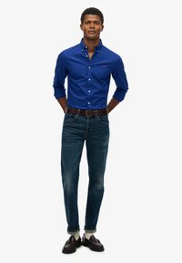 Blauwe button-down overhemD met een logo, gecombineerd met donkere jeans, een bruine riem en instappers, met een gladde textuur en klassieke pasvorm.