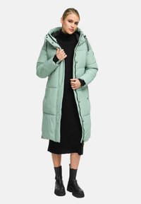 Mintgrön pufferjacka med huva, knapprad framtill, sidofickor och quiltad design; bärs över en svart stickad klänning och svarta stövlar.