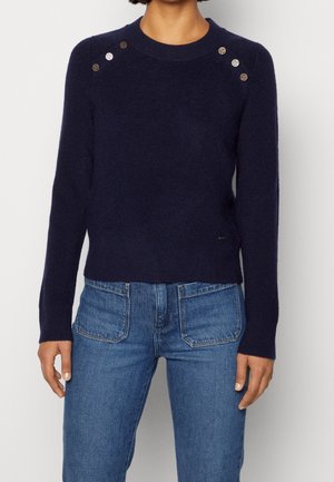 Vrouw die een marineblauwe trui met lange mouwen draagt, voorzien van metalen knopen op de schouders, en een blauwe spijkerbroek.