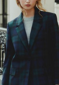 Blazer cu carouri verzi și bleumarin, cu o croială ajustată, rever ascuțit și închidere cu un singur nasture. Purtați peste un pulover gri cu gât rotund.
