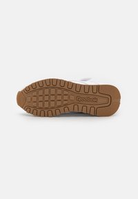 Chaussure de sport blanche avec une semelle en caoutchouc brun texturé, présentant un motif en grille et le nom de la marque "Reebok" embossé sur le côté.