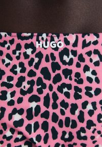 Tkanina s leopardjim vzorom, ki vsebuje črne pike na rožnati podlagi, z belim logotipom "HUGO", postavljenim na vrhu v sredini.