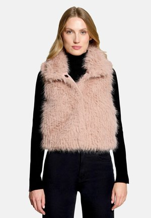 Donna che indossa un gilet soffice rosa chiaro sopra una maglia nera a maniche lunghe e pantaloni neri su uno sfondo bianco uniforme.