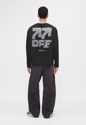 Homme portant une chemise noire à manches longues avec un motif argenté brillant et un texte dans le dos, associée à un jean ample de couleur foncée et des baskets noires.
