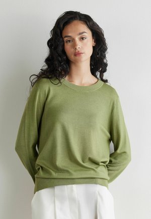 Kvinde med langt, krøllet, mørkt hår iført en løs olivengrøn sweater og hvide bukser, stående foran en ensfarvet lys baggrund.