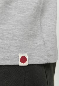 Camiseta de algodón gris con una pequeña etiqueta blanca con un punto rojo en el dobladillo. Textura suave y diseño sencillo con un ajuste clásico.