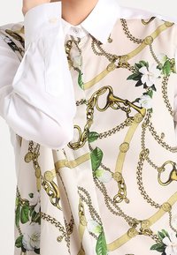 Långärmad skjorta med vit krage; har ett blommigt och guldkedjemönster på en ljus krämfärgad bakgrund. Mjuk tygtextur.