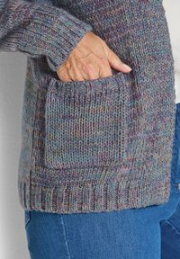 Main dans la poche avant d'un cardigan tricoté multicolore porté sur un haut blanc et un jean bleu.