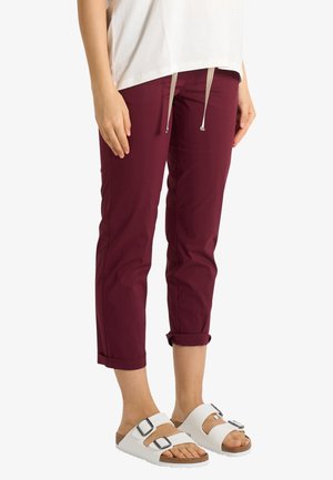 Persona che indossa pantaloni bordeaux risvoltati e sandali bianchi con fibbie, in piedi contro uno sfondo bianco uniforme.