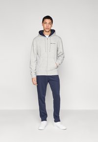 Sweat à capuche gris à fermeture éclair avec intérieur et cordons bleu marine, assorti à un pantalon de jogging bleu marine et des baskets blanches, présenté sur un fond uni.