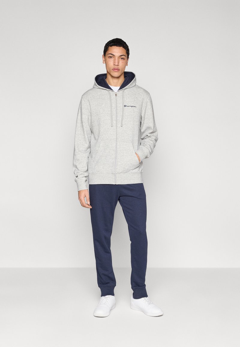 Sweat à capuche gris à fermeture éclair avec intérieur et cordons bleu marine, assorti à un pantalon de jogging bleu marine et des baskets blanches, présenté sur un fond uni.