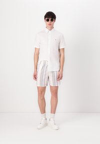 Hvit kortermet skjorte med knapper, stripete lyse bukser med blå og pastellfarger, hvite sneakers og grå solbriller.