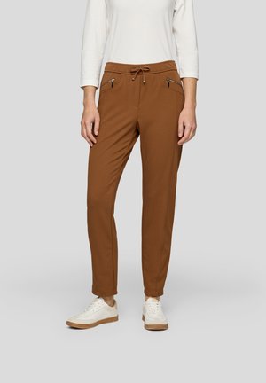 Brown jogger pants with a drawstring waist, two zippered side pockets, and tapered fit. Made from soft, stretchy fabric. Paired with white sneakers.