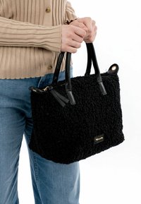 Borsa nera testurizzata con corpo in pelliccia sintetica e manici in pelle nera liscia. Presenta una piccola placca di branding dorata e un dettaglio di nappina.
