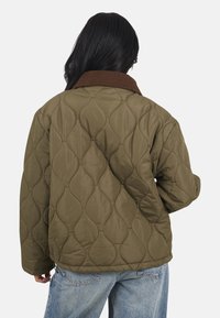 Veste matelassée vert olive avec un col en velours côtelé brun, présentant une coupe ample et un extérieur texturé et à motifs.