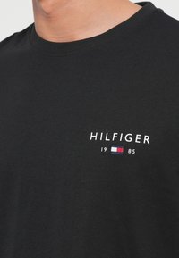 Μαύρο βαμβακερό μπλουζάκι με κανονικό λαιμό, κεντημένο λογότυπο "HILFIGER" και λεπτομέρεια με χρωματικό μπλοκ σε κόκκινο, λευκό και μπλε.