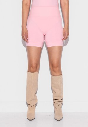 Person iført høytlivede rosa biker-shorts og beige slappe knelange støvler, stående mot en ensfarget hvit bakgrunn.