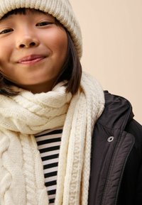 Adīta krēma krāsa cepure un šalle ar kabeli dizainiem, kas valkāta pār melnu jaku un melnu un baltu svītrainu blūzi, ar teksturētām audumiem.