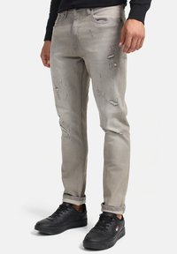 Jeans de mezclilla gris con detalles desgastados, corte slim y costuras visibles. Combinados con zapatillas negras y puños doblados.