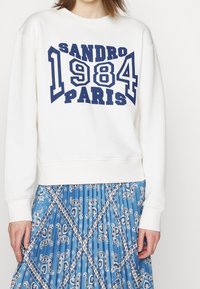 Valkoinen huppari, jossa on tummansininen graafinen teksti "SANDRO 1984 PARIS" edessä; yhdistetty siniseen kuvioituun hameeseen, jossa on paisley-kuvioita.