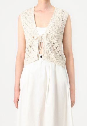 Veste sans manches beige en tricot avec motif en losange, nouée à l'avant, portée par-dessus un haut blanc court et une jupe blanche taille haute.