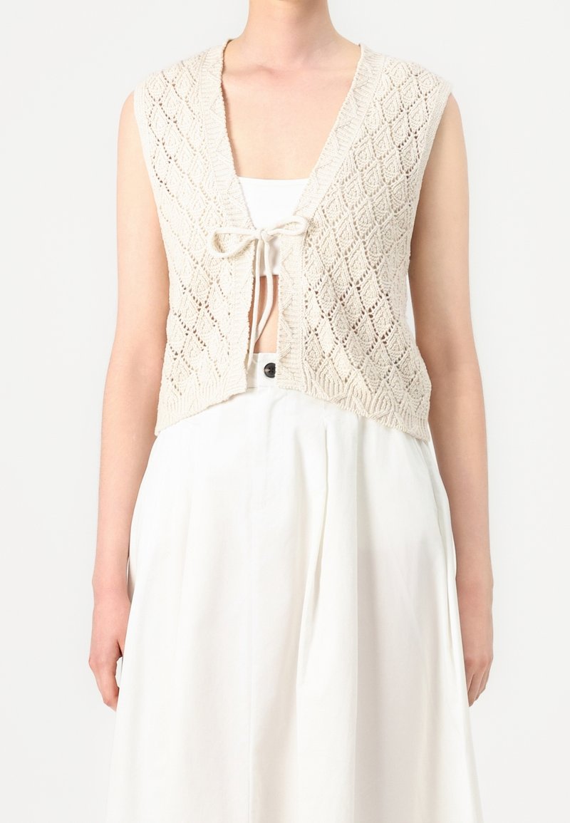 Veste sans manches beige en tricot avec motif en losange, nouée à l'avant, portée par-dessus un haut blanc court et une jupe blanche taille haute.