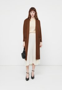 Cardigan lungo marrone in maglina testurizzata con due tasche anteriori, indossato sopra una maglietta beige chiara e una gonna plissè crema, abbinato a tacchi neri.