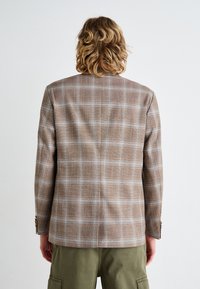 Brown plaid blazer with a structured silhouette. Textured fabric features a mix of brown and cream lines. Single vent at the back, buttoned cuffs.