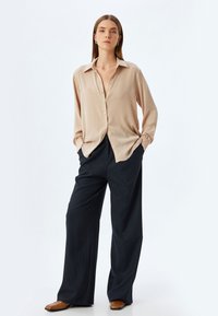 Camicia beige con colletto, abbinata a pantaloni larghi a righe verticali blu scuro. Modello che indossa scarpe slip-on marroni.