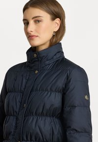 Sötétkék pufferszék, párnázott dizájnnal, magas gallérral és arany színű fémrészekkel. Logó a bal ujján. Simaszerkezet.