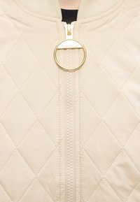 Μπεζ τζάκετ με ραφές σε σχέδιο διαμαντιού και χρυσό στρογγυλό φερμουάρ στο γιακά, χαραγμένο με τη λέξη "Barbour".