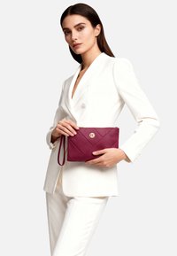 Pochette in pelle color bordeaux con motivo trapuntato diagonale, dettagli dorati e cinturino da polso. La modella indossa un completo bianco per creare contrasto.