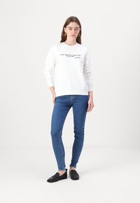 Felpa bianca con testo, polsini e orlo a coste. Abbinata a jeans skinny blu e scarpe slip-on nere. Outfit semplice e casual.