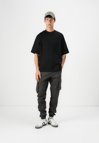 T-shirt nera a girocollo, vestibilità ampia; pantaloni cargo grigi con tasche laterali; sneakers bianche con strisce nere; cappellino beige. Sfondo semplice.