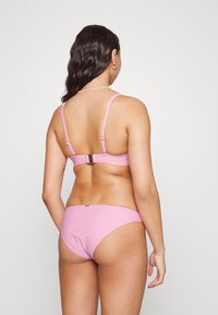 Biquíni rosa com tiras finas e fecho nas costas. Textura suave com design minimalista, apresentando um pequeno detalhe de logótipo na parte traseira.