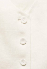 Top en lin blanc avec un décolleté en V et trois boutons ronds recouverts de tissu le long de la patte de boutonnage. Surface texturée avec des coutures subtiles.