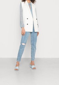 Blazer blanc sans manches à double boutonnière, blouse bleue à motifs floraux, jean déchiré bleu clair, et sandales à talons carrés bleu clair.