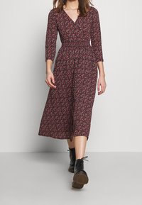 Langärmliges, schwarzes Kleid mit floralem Muster in rot und weiß. Verfügt über einen V-Ausschnitt und eine taillierte Taille, die bis zur Mitte der Waden reicht.