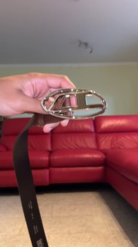 Μαύρη δερμάτινη ζώνη με γυαλιστερή ασημένια οβάλ αγκράφα που φέρει λογότυπο με κοπή. Το φόντο περιλαμβάνει έναν κόκκινο καναπέ.