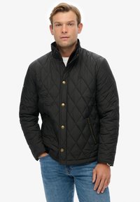 Zwarte gewatteerde jas met een hoge kraag, goudkleurige knoopsluitingen, met rits gesloten zakken en een gladde afwerking. Draagt over een wit shirt.