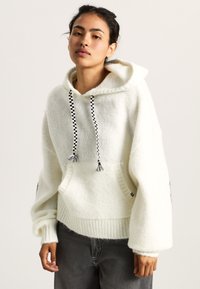 Sweater bianco con cappuccio realizzato in un morbido tessuto testurizzato. Presenta lacci a quadretti neri e una grande tasca frontale con orlo a costine.
