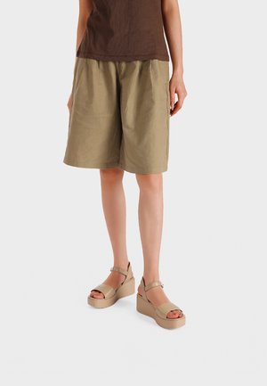 Persona che indossa pantaloncini kaki fino al ginocchio, top marrone e sandali platform beige con cinturini alla caviglia, in piedi su sfondo bianco.