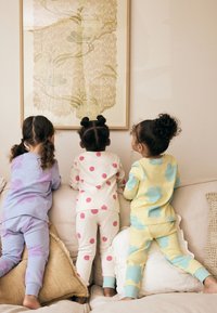 Trois enfants en pyjama : lavande avec des nuages, crème avec des points roses, et jaune avec des fleurs bleu sarcelle, tous faisant face à une œuvre d'art murale.